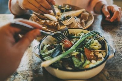 Two people are eating a meal, with one hand holding a fork over a bowl of salad containing greens and vegetables, and another plate of food visible in the background.