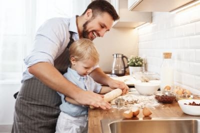 Man and young child roll out dough together on a kitchen counter with baking ingredients and utensils visible around them.
